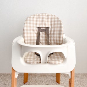 K&ouml;nnte beinhalten: Ein wei&szlig;er Hochstuhl mit einer beige-wei&szlig;en Gingham-Muster-Bezug. Der Stuhl hat einen Holzfu&szlig; und ein Tablett.