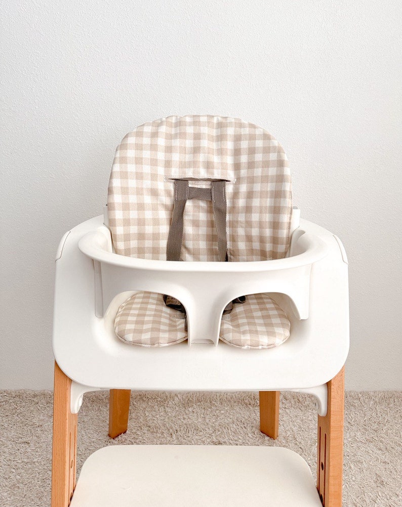 K&ouml;nnte beinhalten: Ein wei&szlig;er Hochstuhl mit einer beige-wei&szlig;en Gingham-Muster-Bezug. Der Stuhl hat einen Holzfu&szlig; und ein Tablett.