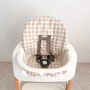 K&ouml;nnte beinhalten: Ein wei&szlig;er Hochstuhl mit einer Sitzbezug in beige-wei&szlig;em Gingham-Muster. Der Stuhl hat eine Holzbasis und einen F&uuml;nf-Punkt-Gurt.