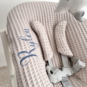 May include: A light beige high chair cover with a waffle weave texture. The cover has blue embroidered text that reads "Rylea". The high chair has gray straps and a warning label. A star-shaped plush toy is attached to the chair.