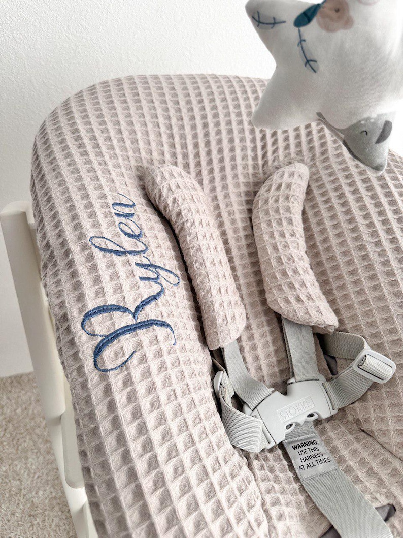 May include: A light beige high chair cover with a waffle weave texture. The cover has blue embroidered text that reads "Rylea". The high chair has gray straps and a warning label. A star-shaped plush toy is attached to the chair.