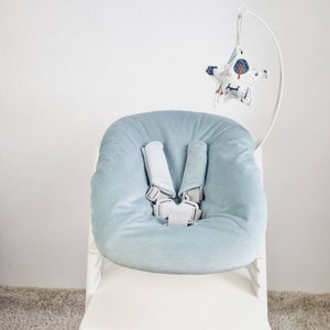May include: A light blue baby seat with a soft, corduroy texture. The seat has a five-point harness with gray straps and a white buckle. A star-shaped mobile with a forest pattern hangs above the seat, attached to a white metal frame.