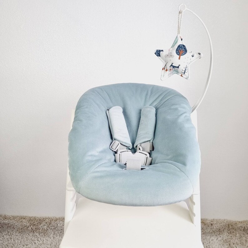 May include: A light blue baby seat with a soft, corduroy texture. The seat has a five-point harness with gray straps and a white buckle. A star-shaped mobile with a forest pattern hangs above the seat, attached to a white metal frame.