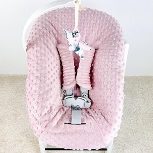 May include: A pink baby seat cover with a textured, dotted pattern. The cover has a gray safety harness and a decorative star-shaped toy hanging from the top. The seat is set on a white frame.