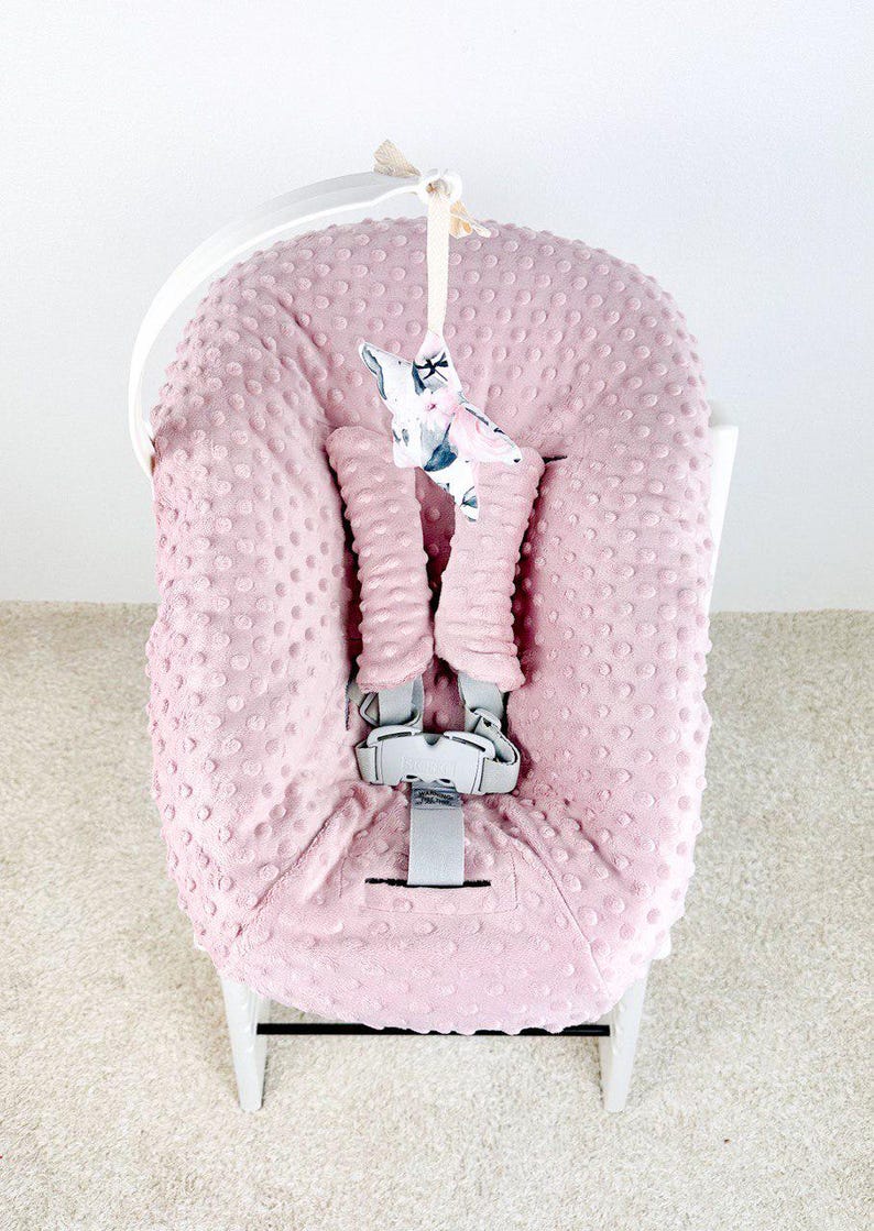 May include: A pink baby seat cover with a textured, dotted pattern. The cover has a gray safety harness and a decorative star-shaped toy hanging from the top. The seat is set on a white frame.