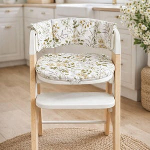 May include: A white high chair with a light wood frame and a floral patterned cushion. The cushion and backrest feature a white background with green and yellow botanical designs. The chair is on a woven jute rug.