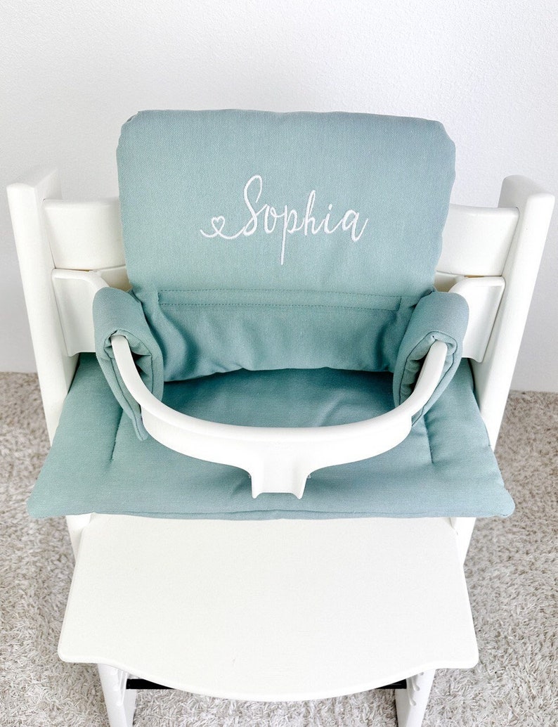 K&ouml;nnte beinhalten: Ein wei&szlig;er Hochstuhl mit einem hellblauen gepolsterten Sitz und R&uuml;ckenlehne. Der Sitz hat einen wei&szlig;en gestickten Namen "Sophia" darauf.