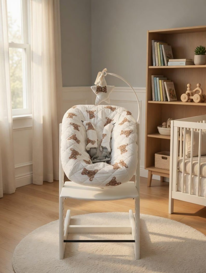 May include: A white high chair with a padded seat featuring a teddy bear pattern. The chair has a safety harness and a mobile with a star and teddy bear. The chair is on a white rug in a room with a bookshelf and crib.
