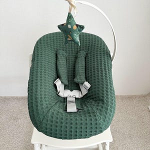 May include: A green waffle-textured baby seat cover with a five-point star-shaped toy hanging above. The seat has a harness and is on a white wooden high chair. The star toy is green with a floral pattern.