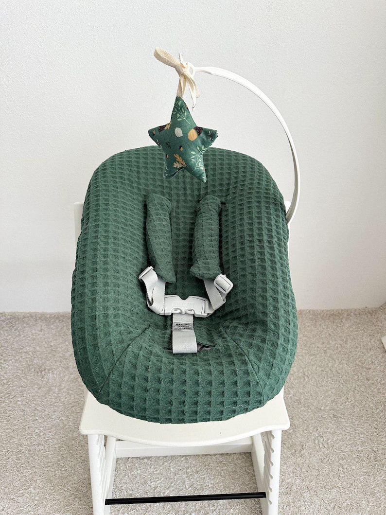 May include: A green waffle-textured baby seat cover with a five-point star-shaped toy hanging above. The seat has a harness and is on a white wooden high chair. The star toy is green with a floral pattern.