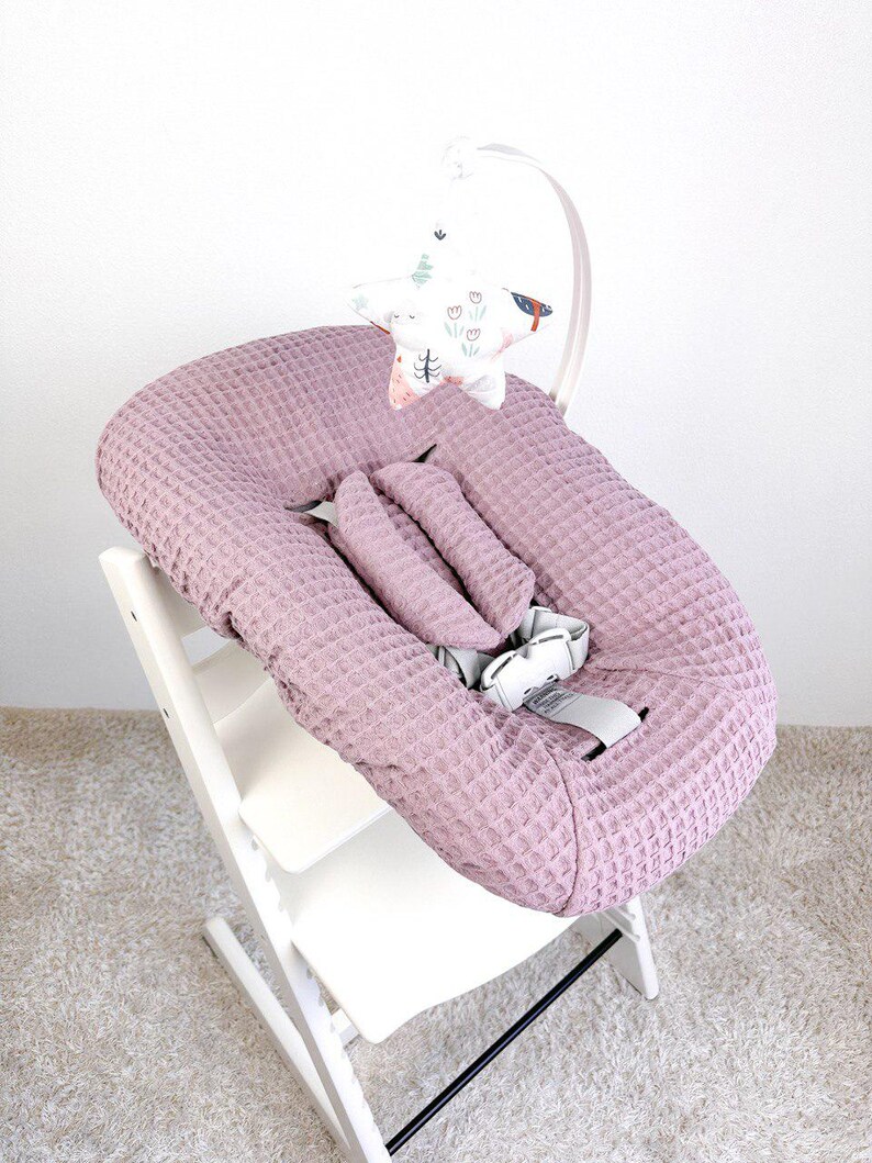 May include: A light purple baby seat with a textured waffle weave fabric. The seat is on a white wooden high chair frame. A decorative mobile with a star-shaped design and floral and bird patterns hangs above the seat.