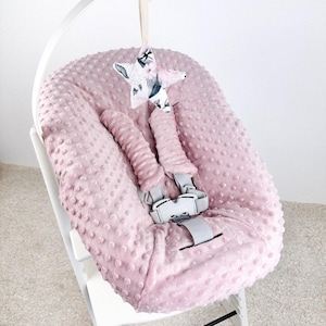 May include: A pink baby high chair with a soft, textured seat cover. The chair has a white frame and gray safety straps. A star-shaped mobile with a floral and animal print hangs above the seat, adding a decorative touch to the nursery.