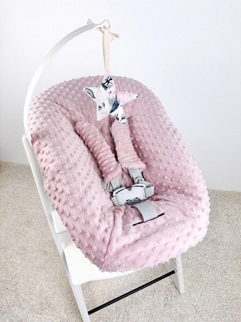 May include: A pink baby high chair with a soft, textured seat cover. The chair has a white frame and gray safety straps. A star-shaped mobile with a floral and animal print hangs above the seat, adding a decorative touch to the nursery.