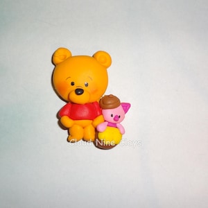 May include: A yellow bear figurine wearing a red shirt standing next to a small pink piglet figurine wearing a yellow shirt and a brown hat. The text Cloud Nine Clays is printed in white across the bottom of the two figures.