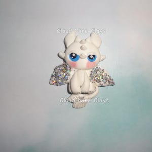 May include: A white dragon figurine with blue eyes, pink cheeks, and glittery wings. The figurine has the text "Cloud Nine Clays" in white. The dragon is in a sitting position.