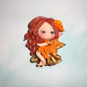 May include: A clay figurine of a character with long wavy auburn hair, large eyes, and pointed ears. The figure wears an orange leaf-shaped top with gold glitter accents and a dark gold skirt. The text Cloud Nine Clays appears twice.