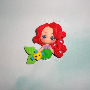 May include: A clay figurine of a mermaid with bright red hair, large blue eyes, and a green tail. The mermaid wears a purple seashell top and holds a small yellow and blue fish. The text Cloud Nine Clays is visible on the figurine.