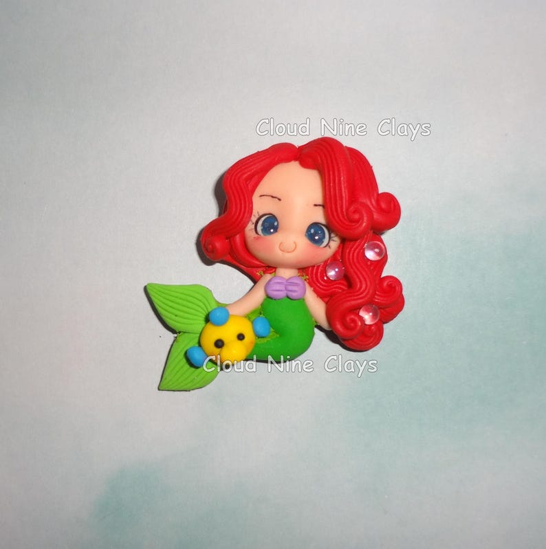 May include: A clay figurine of a mermaid with bright red hair, large blue eyes, and a green tail. The mermaid wears a purple seashell top and holds a small yellow and blue fish. The text Cloud Nine Clays is visible on the figurine.