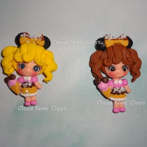 May include: Two clay figurines of characters with large eyes, wearing yellow or brown curly hair, black mouse ears, and bows. Each figure holds a pink and brown ice cream cone, wears a pink top with a brown skirt, and has the text Cloud Nine Clays.
