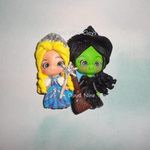 May include: A whimsical figurine featuring a princess with a silver crown and a witch with a black hat. The princess wears a blue dress and holds a wand. The witch has green skin and a black dress, holding a broom. The text "Cloud Nine Clays" is visible.