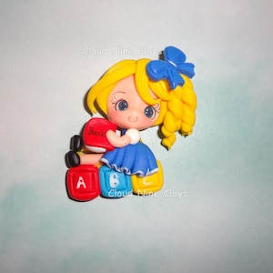 May include: A clay figurine of a character with yellow hair and a blue bow, wearing a blue dress. The character holds a red book labeled Book and sits atop three square blocks colored red, blue, and yellow, marked with the letters A, B, and C.