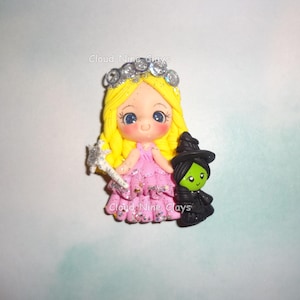 May include: A handcrafted polymer clay figurine featuring a princess with a silver crown, blonde hair, and a pink dress holding a wand. A small green-skinned witch in a black hat and dress stands beside her. The text "Cloud Nine Clays" is visible.