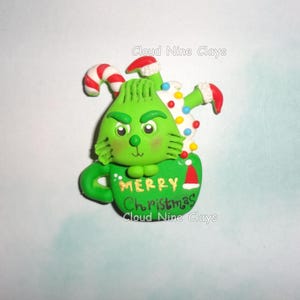 May include: A green clay figure with a grumpy expression sits inside a green mug labeled Merry Christmas. The figure is surrounded by a red and white candy cane, a white whipped cream topping with colorful sprinkles, and a small red Santa hat.