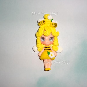 May include: A handcrafted polymer clay figurine of a bee fairy. The fairy has yellow hair, a yellow dress with black stripes, and white wings. The figurine has a flower on the dress and two bees on top of the head. The text "Cloud Nine Clays" is visible.