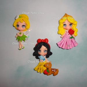 May include: Three handmade clay figurines of fairytale characters. One is a fairy in a green dress with wings, another is a princess in a pink dress holding a rose, and the third is a princess in a yellow dress with a deer.