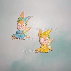 May include: Two clay figurines of bunny fairies. One wears a blue dress and flower crown, the other a yellow dress and flower crown. Both have closed eyes, pink cheeks, and iridescent wings. The text "Cloud Nine Clays" is visible.