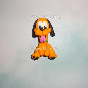 May include: A handcrafted orange dog figurine with black ears and a pink tongue. The dog has a green collar and black paws. The text "Cloud Nine Clays" is visible on the image.