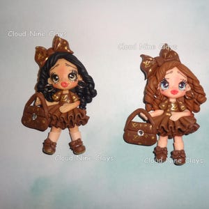 May include: Two clay figurines of girls with large eyes, wearing brown dresses with gold patterns, matching bows, and carrying brown handbags with a central rhinestone. One figure has black hair, the other has brown hair. Text reads Cloud Nine Clays.