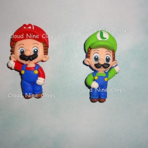 May include: Two handmade clay figurines of video game characters Mario and Luigi. Mario wears a red hat, red shirt, blue overalls, and has a black mustache. Luigi wears a green hat, green shirt, blue overalls, and a black mustache. The text "Cloud Nine Clays" is visible.