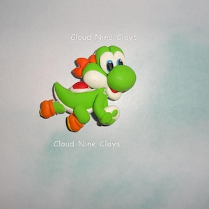 May include: A green clay figurine of a dinosaur character with large white eyes, orange shoes, and a red saddle on its back. The figure has a white snout and belly. The text Cloud Nine Clays appears twice in the image, once above and once below.