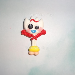 May include: A white clay character figurine with a red arched eyebrow, mismatched black eyes, blue lips, and pink cheeks. It has red arms crossed over its chest, a yellow base, and two small peach-colored feet. The text Cloud Nine Clays is visible.