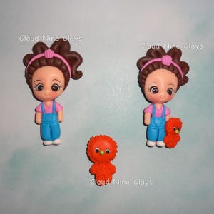 May include: Handmade polymer clay figurines. Two dolls with brown hair, pink shirts, and blue overalls. One doll holds an orange creature. An orange creature is also shown separately. The text "Cloud Nine Clays" is visible.