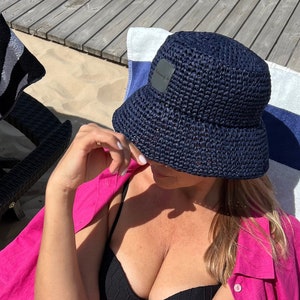 May include: A woman wearing a pink linen shirt, black bikini top, and pink linen shorts with a "SIMPLE" label. She is sitting on a beach chair with a blue woven bucket hat on her head.
