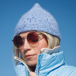 May include: A light blue puffer jacket, a light blue knit beanie, and round sunglasses with brown lenses. The jacket has a zipper closure and a high collar. The beanie has a speckled pattern. The background is a clear, bright blue sky.