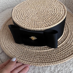 Puede incluir: Un sombrero de paja beige con copa plana y ala ancha. El sombrero presenta una cinta de grosgrain negra ancha envuelta alrededor de la base de la copa, atada en un lazo en la parte delantera. Un pequeño emblema de metal dorado está centrado en el lazo.