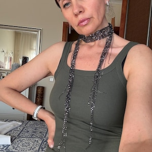 May include: A dark olive green ribbed vest top paired with a long, thin, dark metallic grey scarf featuring a delicate, open-knit, shimmering texture that wraps around the neck and hangs down in two slender, dangling strands.