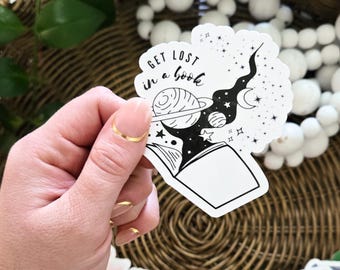 May include: A white sticker with a black and white illustration of a book with a galaxy inside. The text "Get lost in a book" is written above the book.