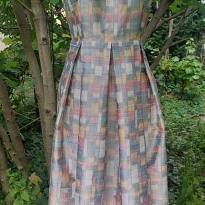 Può includere: Un abito senza maniche con scollo a barchetta e corpetto aderente che si trasforma in una gonna a pieghe. Il tessuto presenta un motivo a griglia multicolore in stile intrecciato nei toni del giallo, rosa, blu e grigio su fondo chiaro.