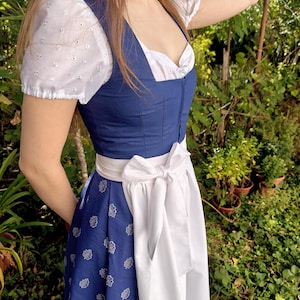 May include: A blue dirndl dress featuring a square neckline and a fitted bodice. The skirt is blue with a white leaf pattern, paired with a white apron tied in a bow at the waist. The blouse is white with short puff sleeves and eyelet embroidery.