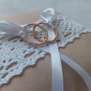 May include: A peach-colored ring pillow with a white lace band and white satin ribbon. Two rose gold wedding rings are tied to the pillow with the ribbon. The pillow is a soft square shape.