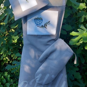 Puede incluir: Delantal de cocina gris con un diseño de rama de olivo bordada en blanco y un guante de horno gris a juego. El delantal tiene un bolsillo grande y el texto "ofili" es visible en la parte inferior.
