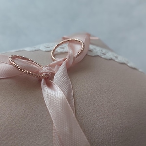May include: A blush pink ring pillow with a delicate lace trim. Two gold rings are secured to the pillow with a matching satin ribbon. The pillow is a soft, textured fabric, perfect for a wedding ceremony.