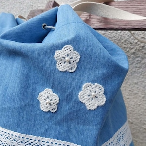 Può includere: Borsa blu in denim con coulisse, rifinita con pizzo bianco e tre applicazioni floreali bianche all'uncinetto. Ha un cinturino color crema. Ideale per trasportare oggetti.