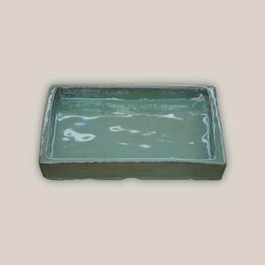 May include: A rectangular, ceramic tray in a muted teal color. The tray has a slightly textured surface and a dark rim. The edges are gently curved, and the overall design is simple and modern. This tray is suitable for serving or display.