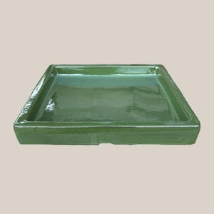 May include: A square, glazed ceramic tray in a vibrant green hue. The tray has a shallow, rectangular interior and slightly raised edges. The glossy finish reflects light, highlighting the smooth surface and clean lines of this decorative piece.