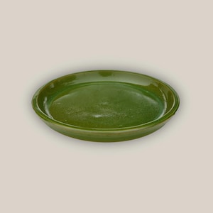 May include: A round, green ceramic dish with a glossy finish. The dish has a slightly raised rim and a smooth, flat interior. The color is a deep, earthy green, suggesting a handmade or rustic aesthetic. The dish is likely used for serving or displaying food.